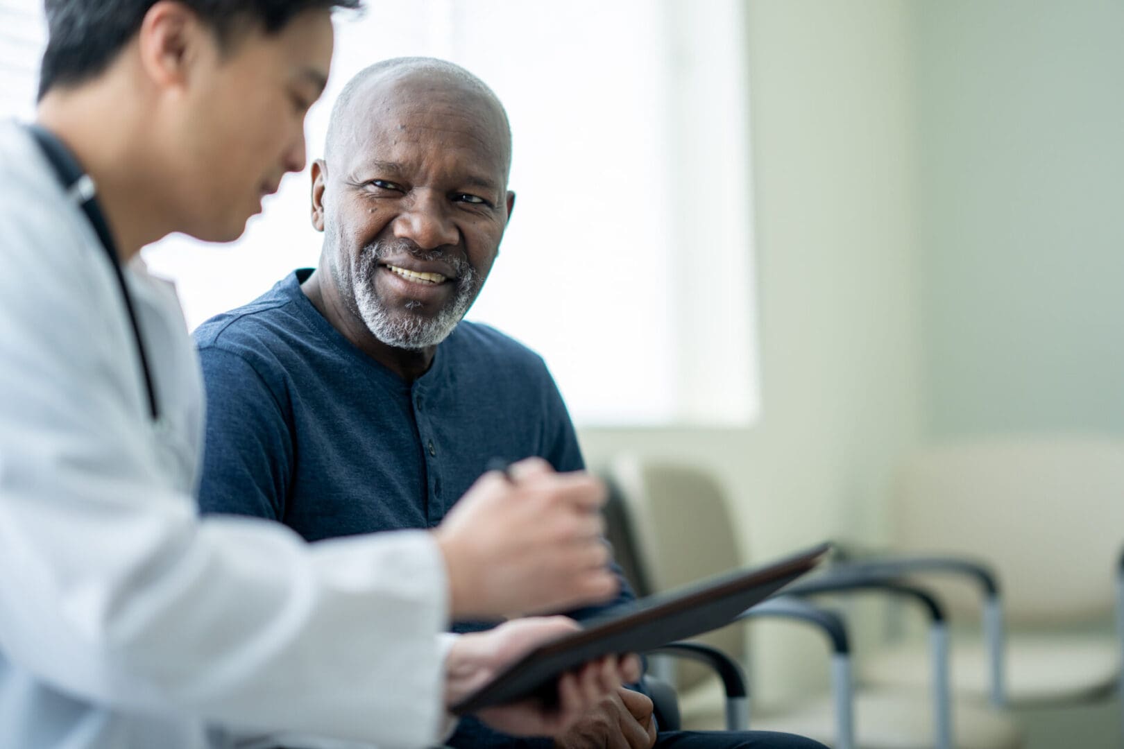 A senior gentleman of African decent sits in a waiting room beside his doctor as they review test results on a tablet together.