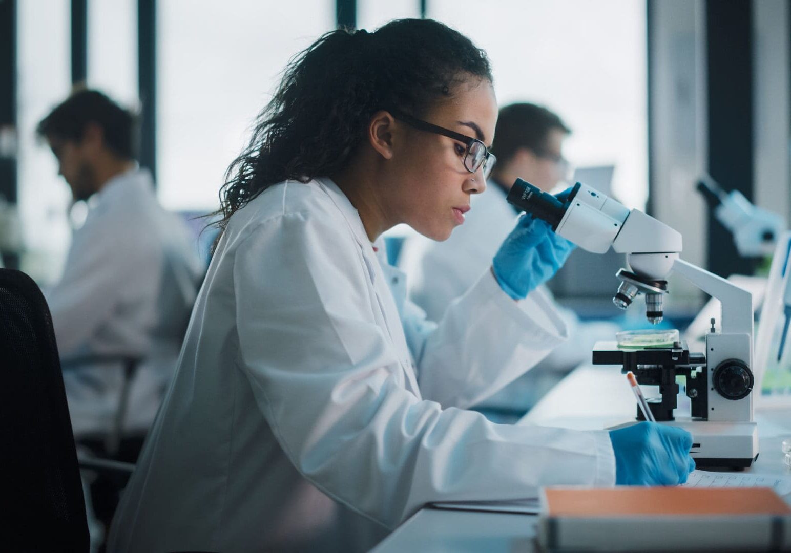 Medical Science Laboratory: Beautiful Black Scientist Looking Under Microscope Does Analysis of Test Sample. Diverse Team of Young Specialists, Using Advanced Technology Equipment.