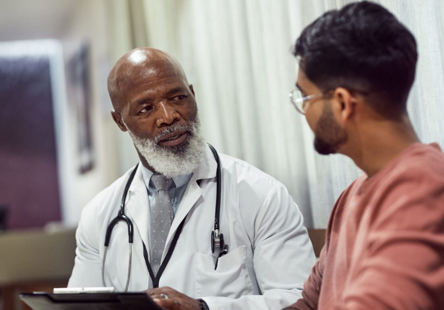 Senior doctor with beard holding clipboard, talking to male patient in emergency room