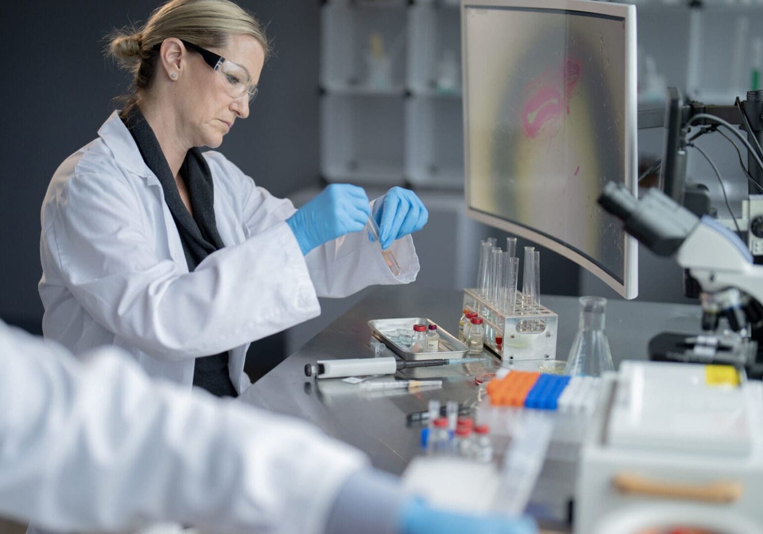 A female medical lab technician is seen working on a vaccine.  She is dressed in a white lab coat and personal protective equipment as she focuses on the task.